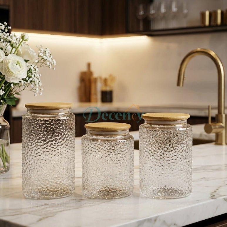 Three airtight clear glass jars with wooden lids arranged on a marble kitchen countertop.