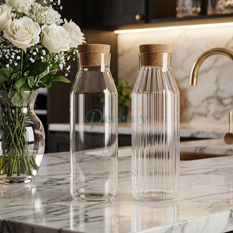 Smooth clear and ribbed fluted 1 litre glass water bottles with cork stoppers on a marble kitchen countertop.
