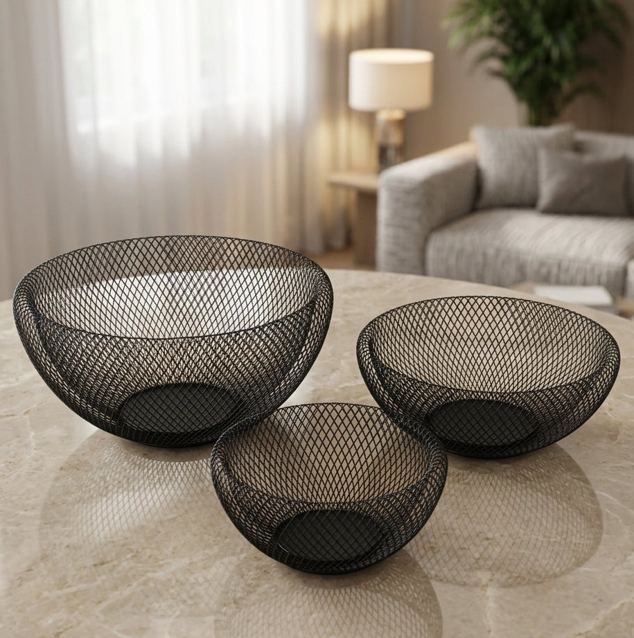 Set of three round black wire mesh metal fruit bowls placed on a marble table in a modern living room.   