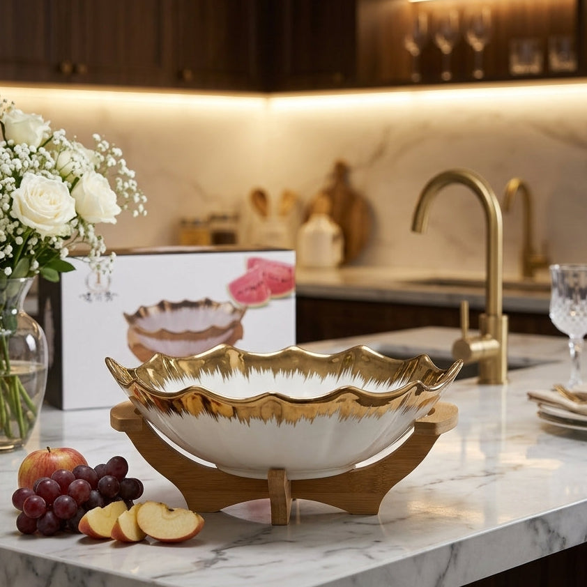 White ceramic fruit bowl with wavy gold crown rim resting on a wooden stand with fresh fruits on a marble kitchen counter.