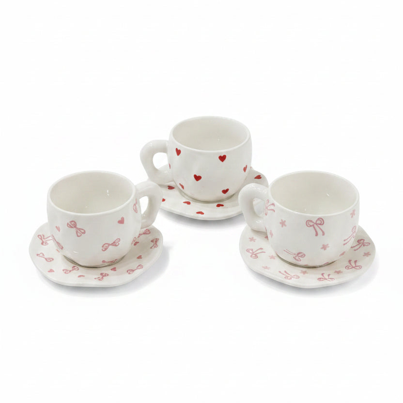 White ceramic tea cups with red heart and pink bow patterns set on saucers over a lace tablecloth.