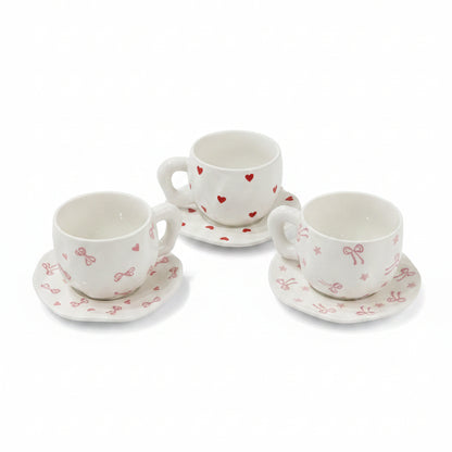 White ceramic tea cups with red heart and pink bow patterns set on saucers over a lace tablecloth.
