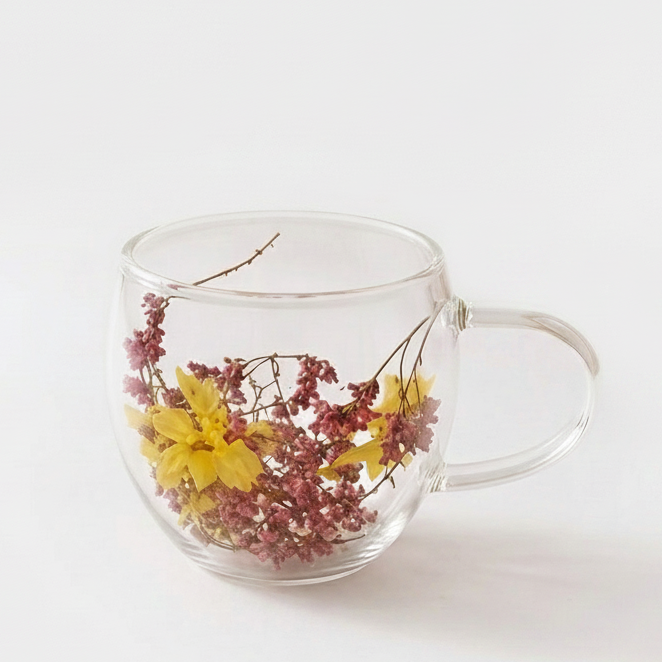 Double wall clear glass coffee mug with pink dried flower petals inside on a woven coaster