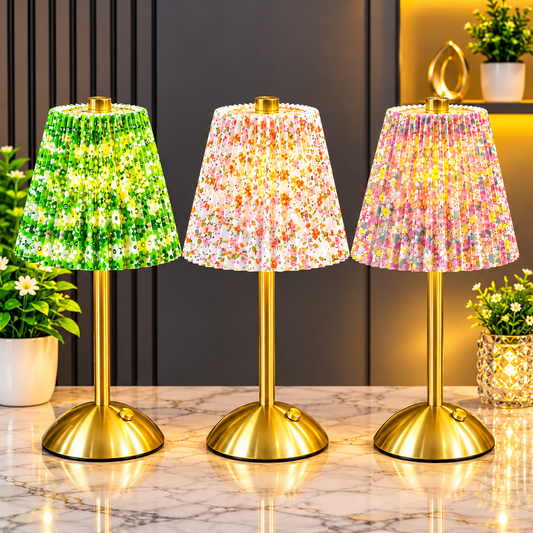 Three table lamps with gold-finished metal bases and pleated fabric shades are arranged on a marble surface. The left lamp has a green floral shade, the center lamp has a pink floral shade, and the right lamp has a multicolor floral shade.