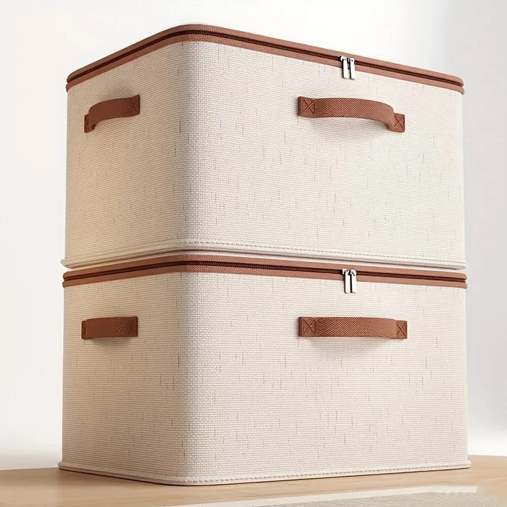 Two stacked beige fabric clothes storage boxes with brown zippers and handles on a wooden table
