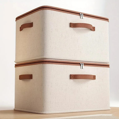 Two stacked beige fabric clothes storage boxes with brown zippers and handles on a wooden table