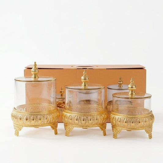 3 piece clear glass jar set with ornate golden metal stands and decorative lids on a plain background