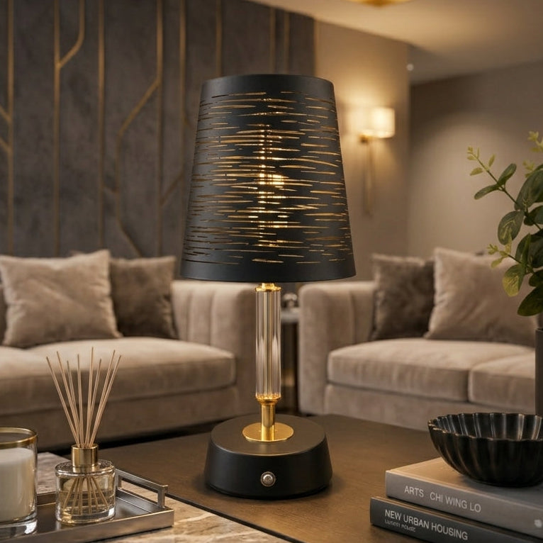 Modern black slitted metal table lamp glowing warm light on a dark wooden side table beside a green plant