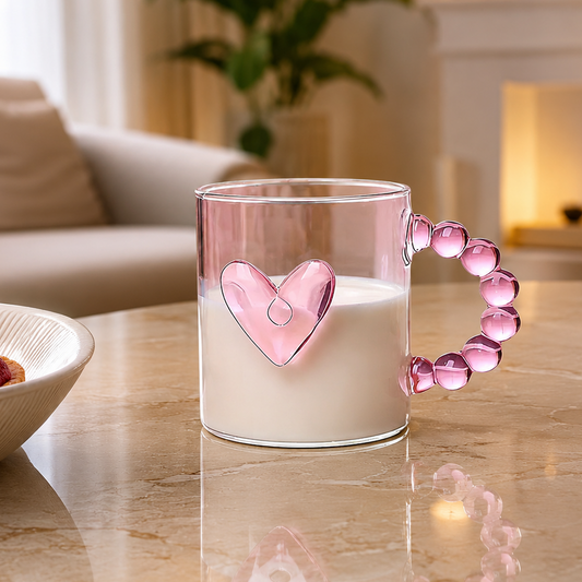 Front view of a clear glass mug featuring a pink heart shape and a bubble-beaded handle filled with milk on a marble table.   