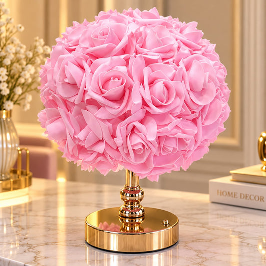 A pink artificial rose ball table lamp on a shiny gold metallic stand, set on a marble surface with a blurred home decor background.