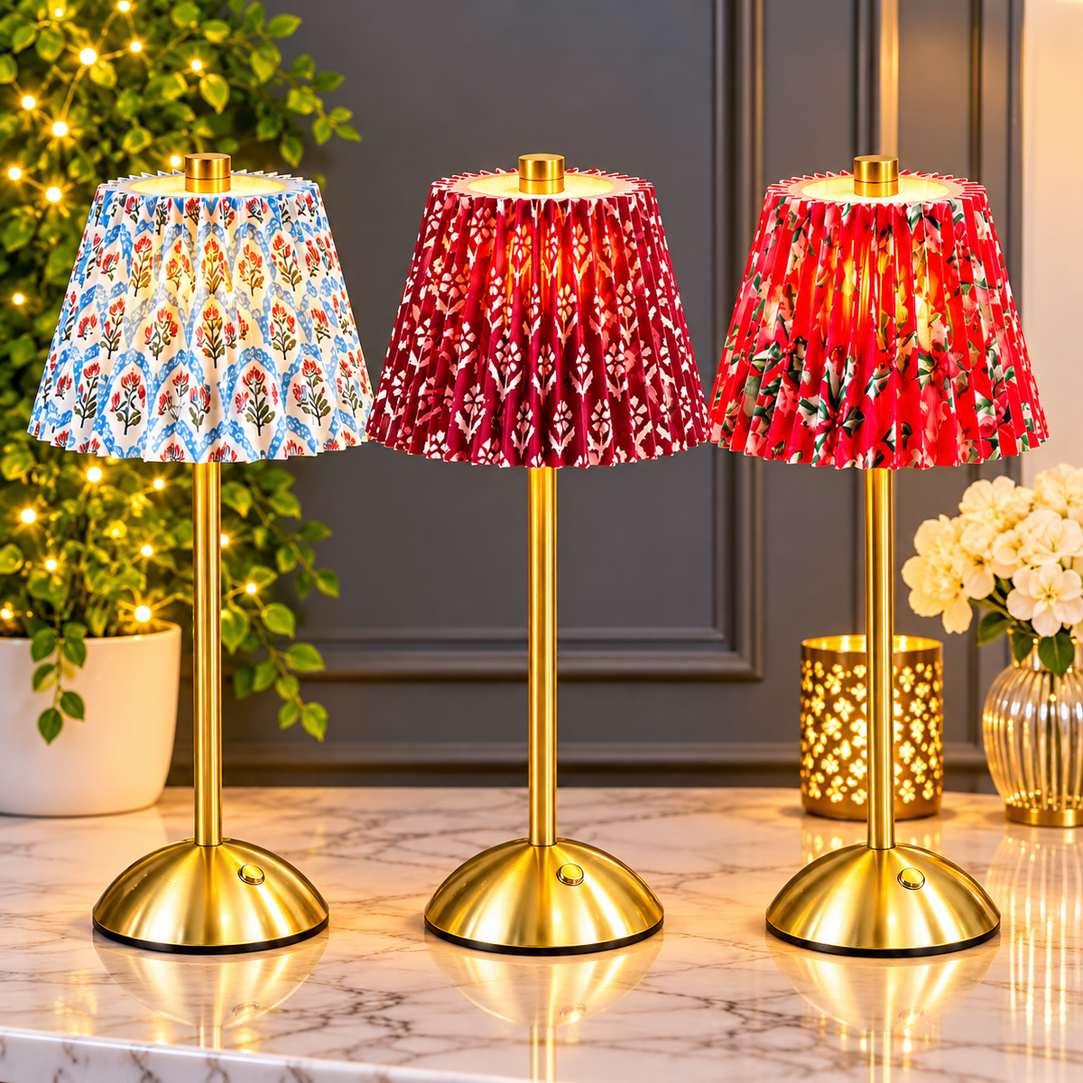 Three decorative table lamps with gold-finished metal bases and pleated fabric shades are arranged on a marble surface. The left lamp has a blue, orange, and white patterned shade, while the center and right lamps have red shades with white floral motifs and small green leaves.