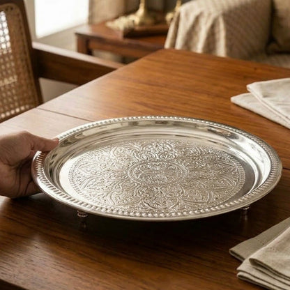 Hand placing a round engraved silver-plated stainless steel platter on a wooden dining table