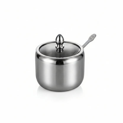 Stainless steel sugar jar container with a clear glass lid and small serving spoon on a marble countertop