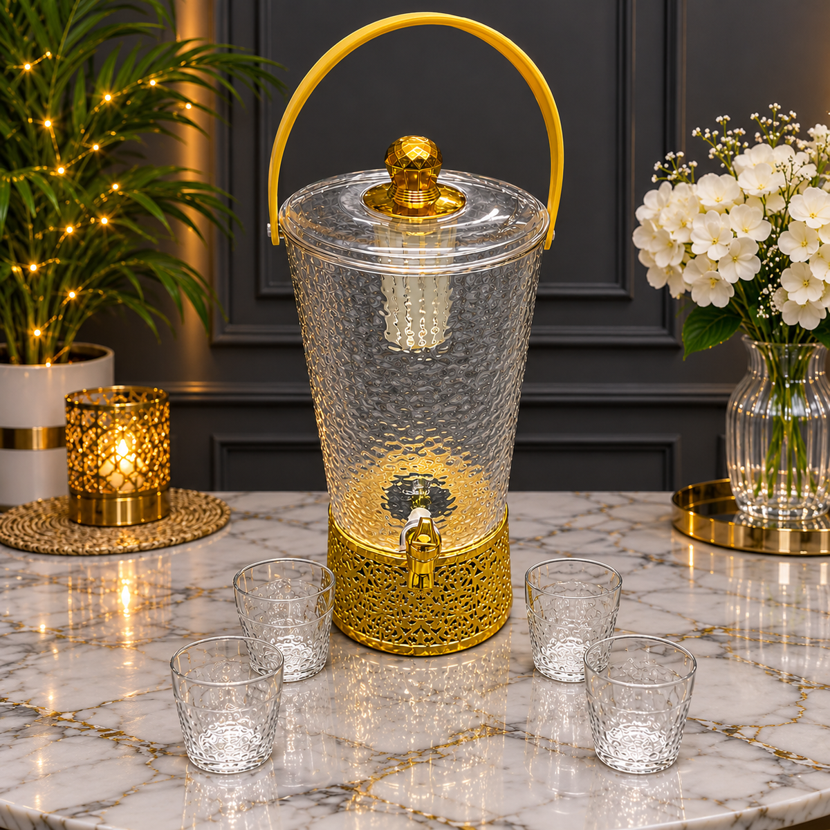 A clear glass water dispenser with a hammered finish, gold-tone metal base and handle, and a small gold spout, shown on a marble countertop with four matching clear glasses. Decorative plants, flowers, and a lit candle are in the background.